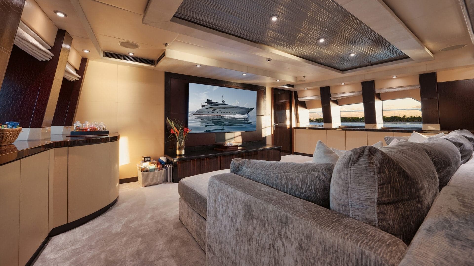 Yacht salon interior with a wall-mounted screen, a gray sectional sofa and warm light coming through the side windows