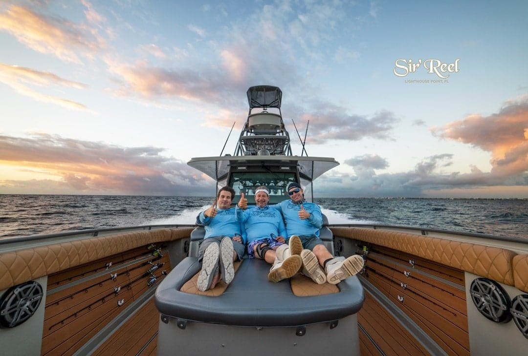 Three people in blue shirts relaxing on the deck of a sportfishing boat at sea with a colorful sunset sky
