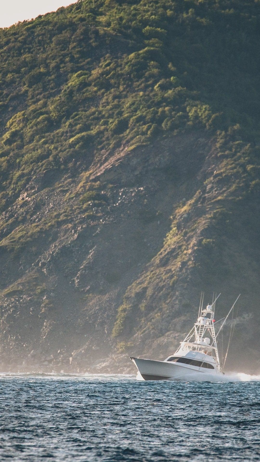 A sportfishing boat cruising near a rugged, green-clad hillside at sea, set against a backdrop of mist and waves