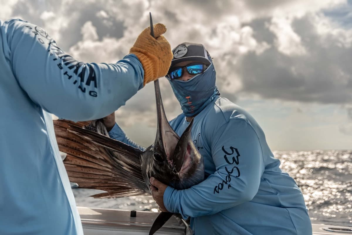 Two fishermen in matching blue gear expertly handle a sailfish on a boat