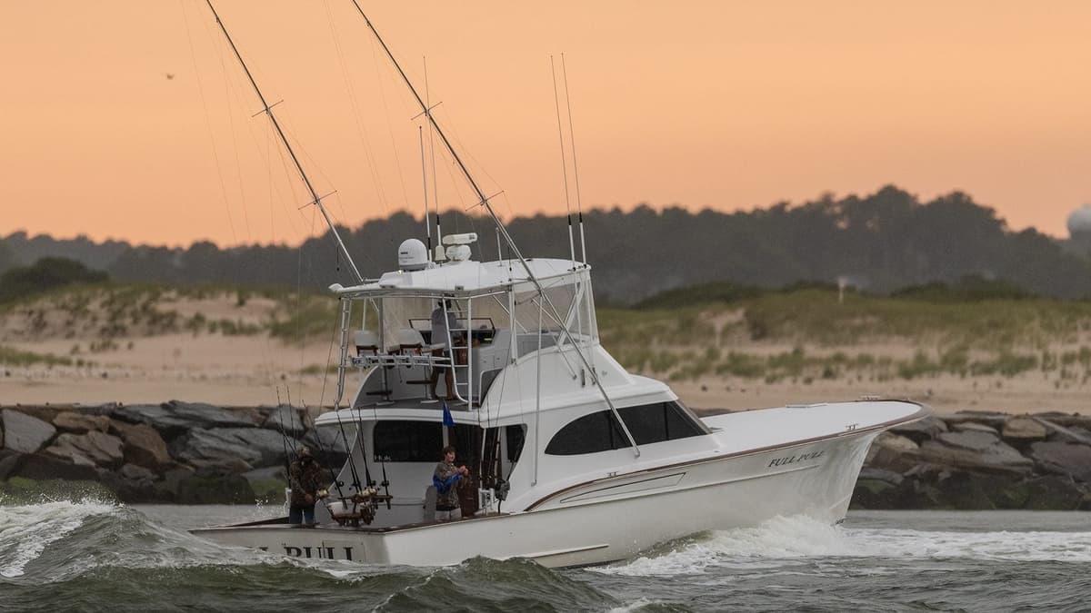 Sportfishing boat navigating through waves with multiple fishing rods, against a backdrop of a sandy coast during sunset