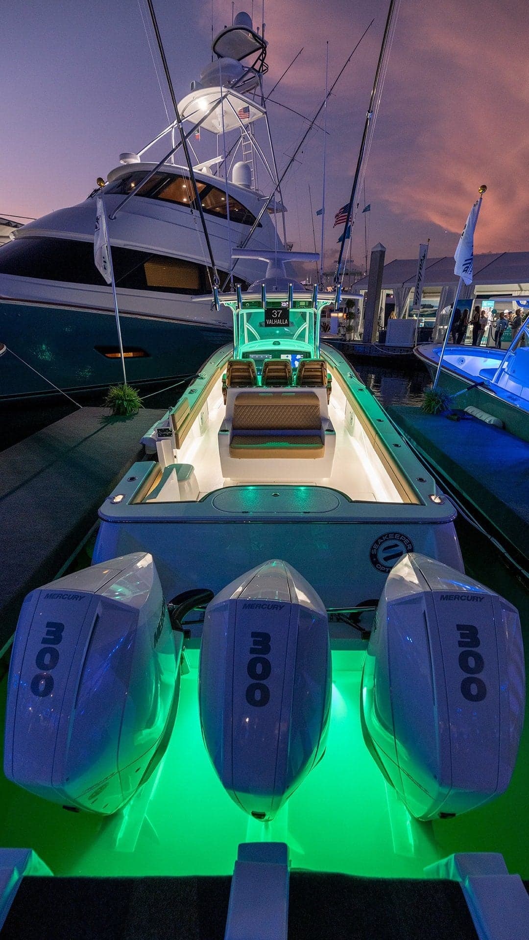 A sleek sportfishing boat docked at sunset, featuring three powerful outboard motors and vibrant green underwater lights