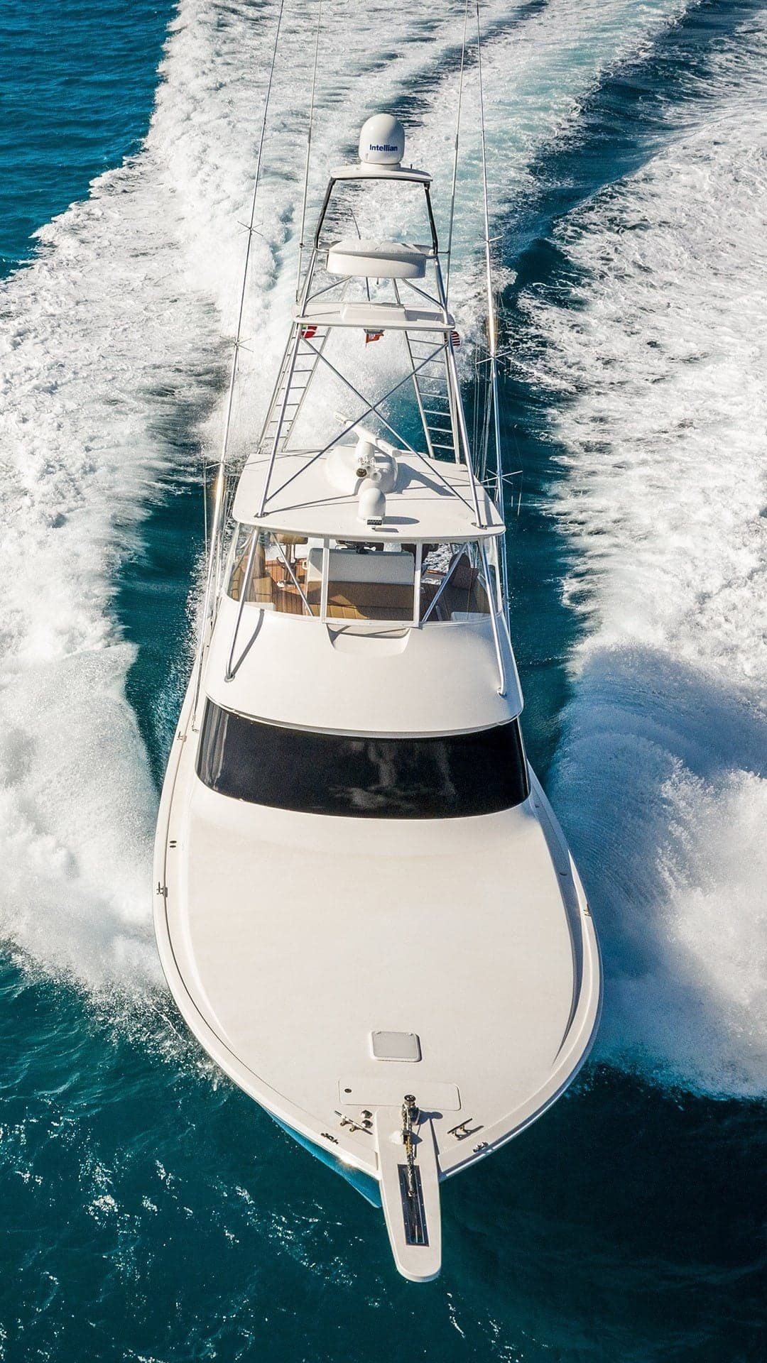 Aerial view of a sleek, white sportfishing yacht cruising at high speed on blue ocean waters, leaving a frothy wake behind