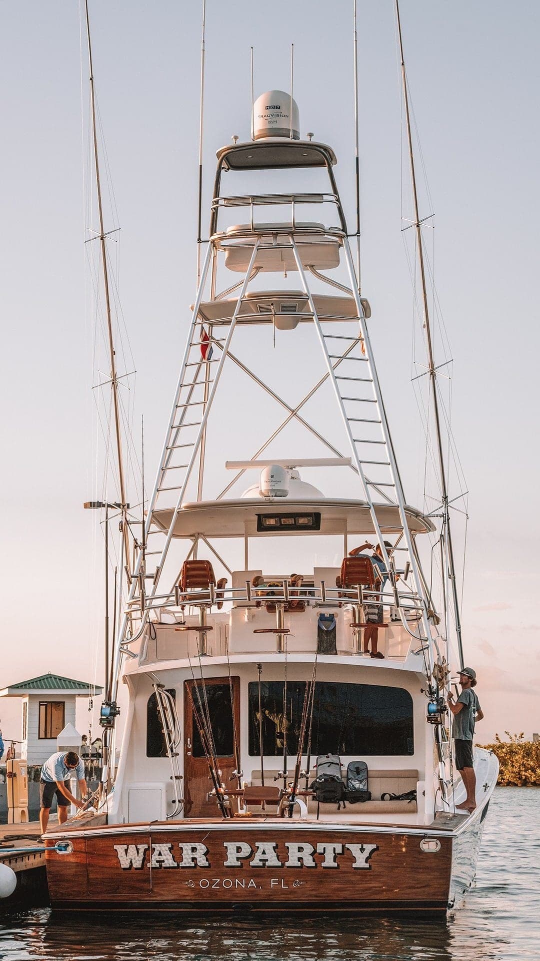 A sportfishing yacht docked at a marina, featuring a towering tuna tower, multiple fishing rods, and a wooden stern